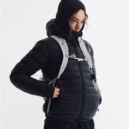 A person wearing an EastLyst Ultra-Light Down Jacket in black with a hood and a light gray backpack stands against a plain white background.