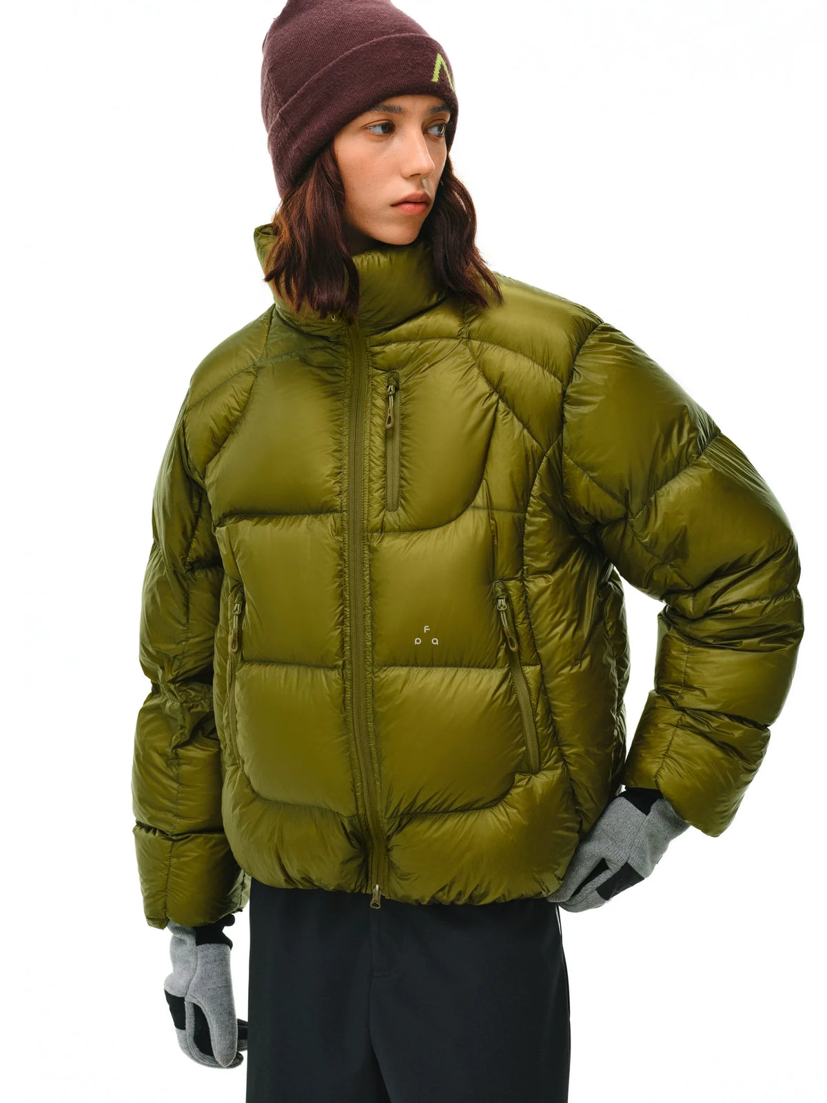 A person in an olive green EastLyst Aurora Puffer Jacket, a burgundy beanie, gray gloves, and black pants stands against a white background, looking to the side with a neutral expression.