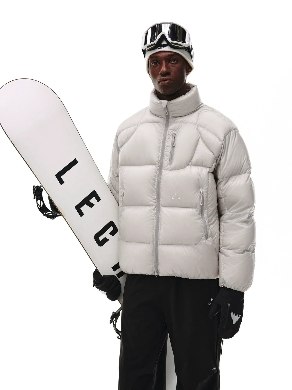 A person in an EastLyst Aurora Puffer Jacket, black snow pants, ski goggles, and gloves stands holding a white snowboard with bold black lettering against a plain white background.