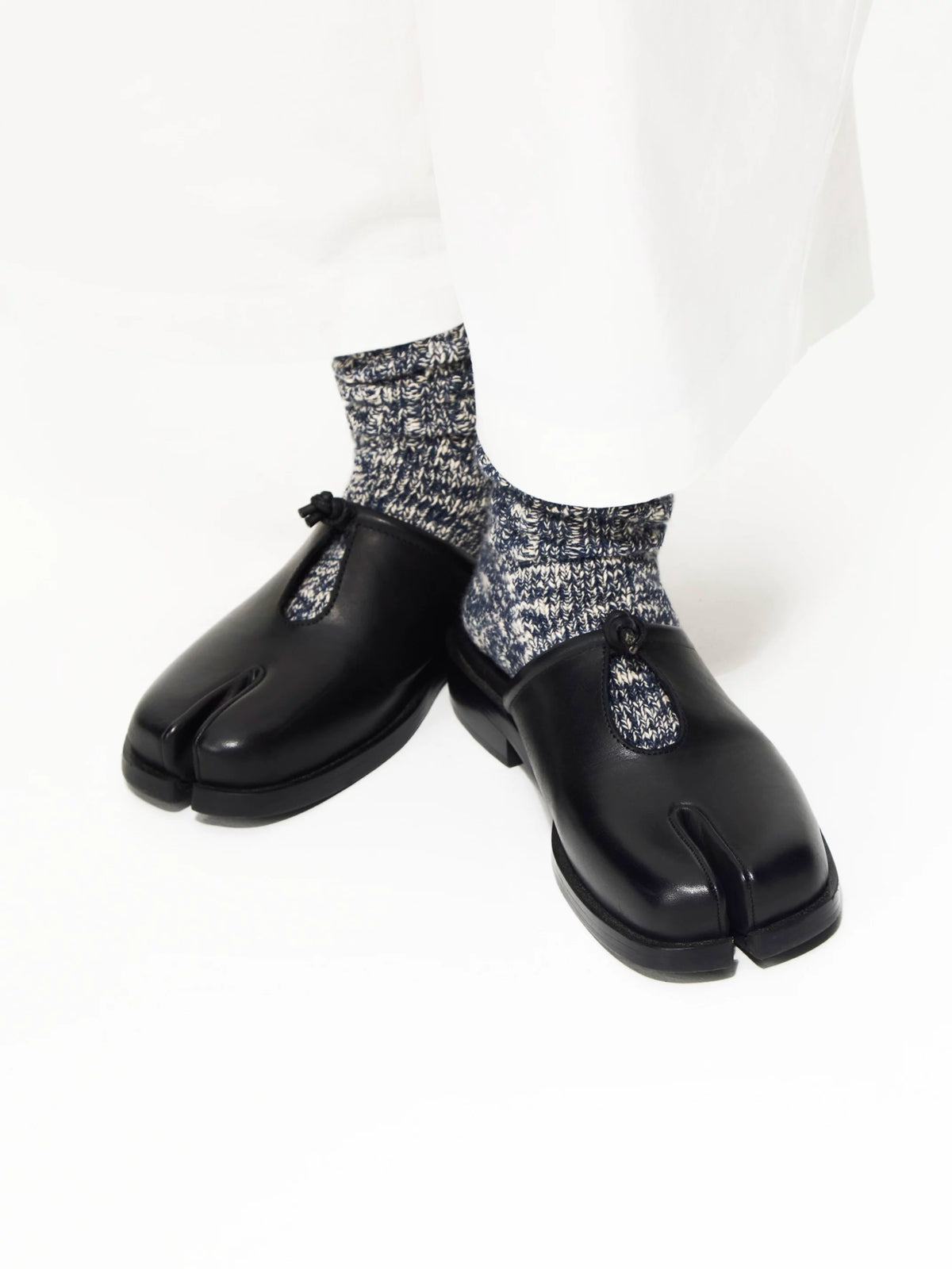 Black split-toe mules with knotted cutout worn with grey speckled socks, front angled view on white background
