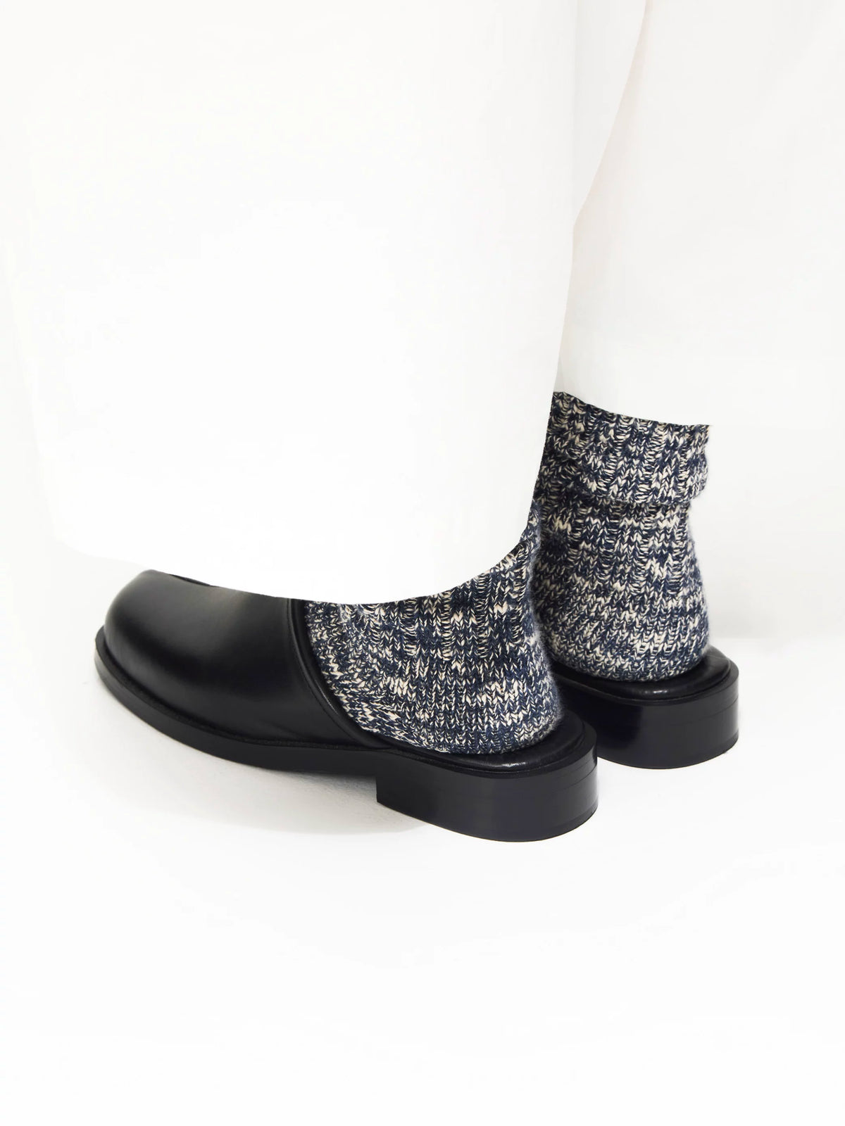 Black leather mules worn with grey speckled socks and white trousers, rear angled view on white background