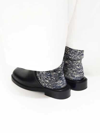 Black leather mules worn with grey speckled socks and white trousers, rear angled view on white background