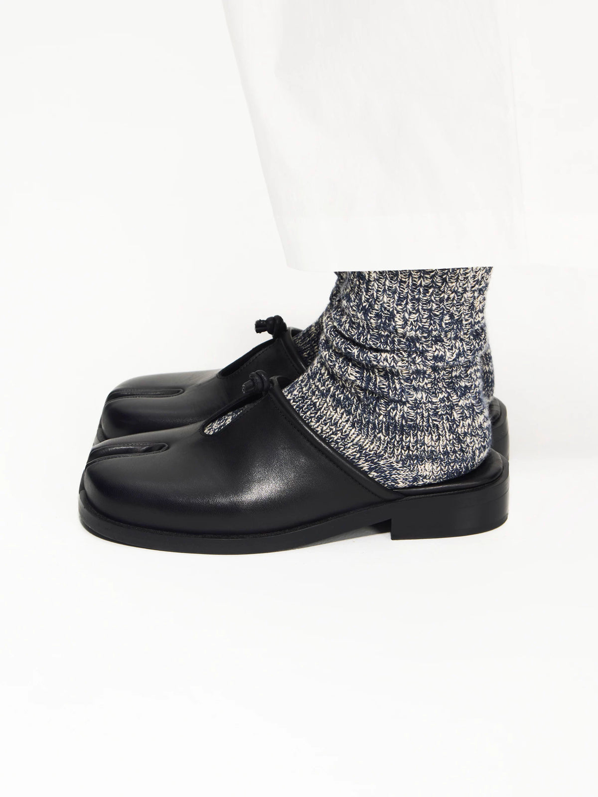 Black split-toe mules worn with grey speckled socks and white trousers, side view on white background