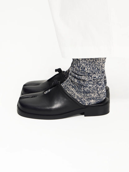 Black split-toe mules worn with grey speckled socks and white trousers, side view on white background