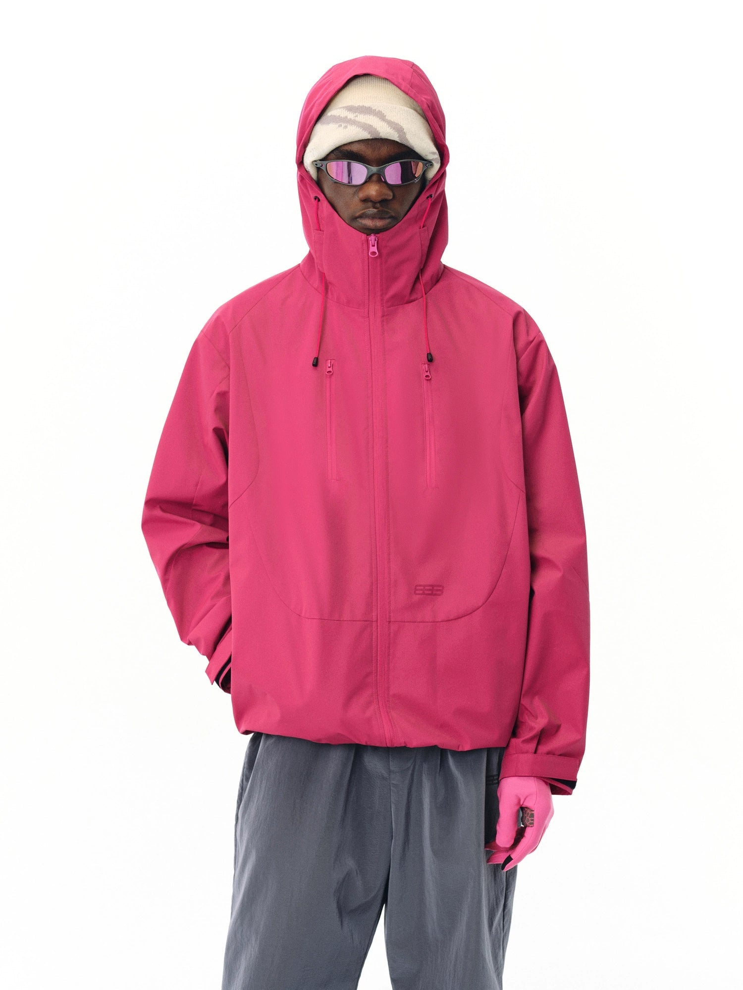 A person wears the EastLyst Best All-Rounder Jacket in bright pink, paired with pink gloves, gray pants, sunglasses, and a beige beanie while standing against a plain white background.