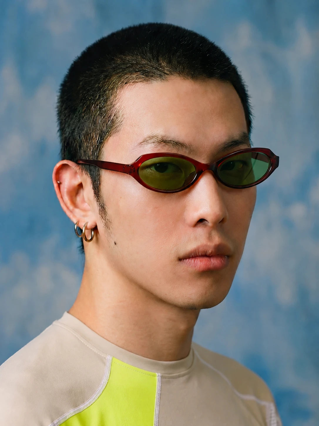 Three-quarter portrait of a model wearing burgundy rounded-square sunglasses with green lenses against a blue background.