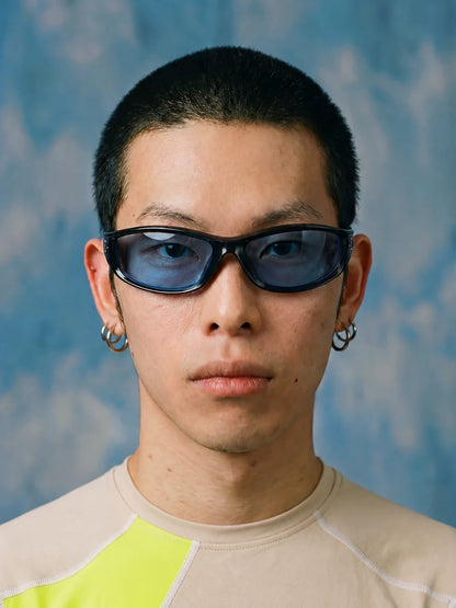 Model wearing black wraparound sunglasses with blue lenses against a blue background.
