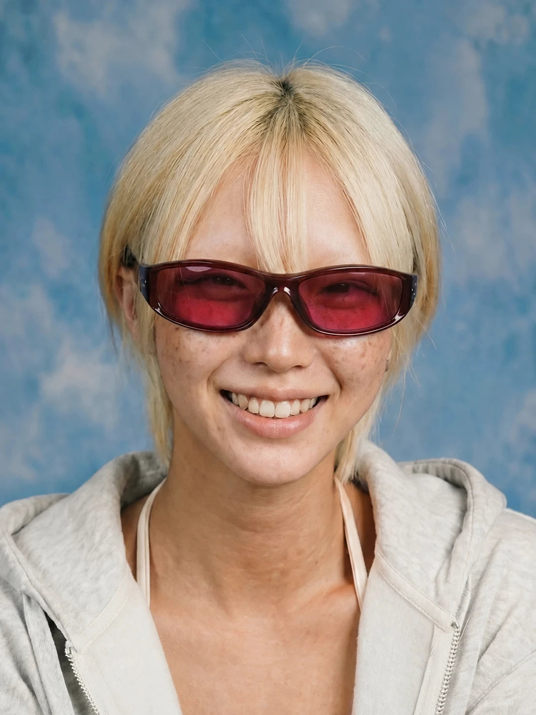Smiling model wearing burgundy wraparound sunglasses with red-tinted lenses and a grey hoodie against a blue background.